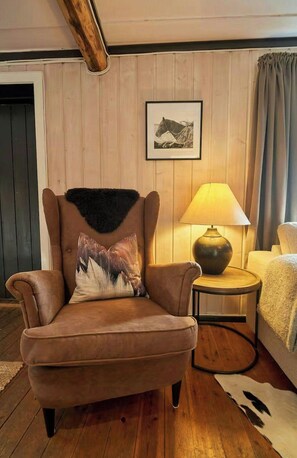 Un sillón de cuero marrón con un plaid de piel y un cojín con diseño de montaña, ubicado junto a un piso de madera y una pequeña mesa redonda con una lámpara.
