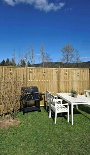 Un jardin avec une clôture en bois, un barbecue, une table avec des chaises et un pot de fleurs.