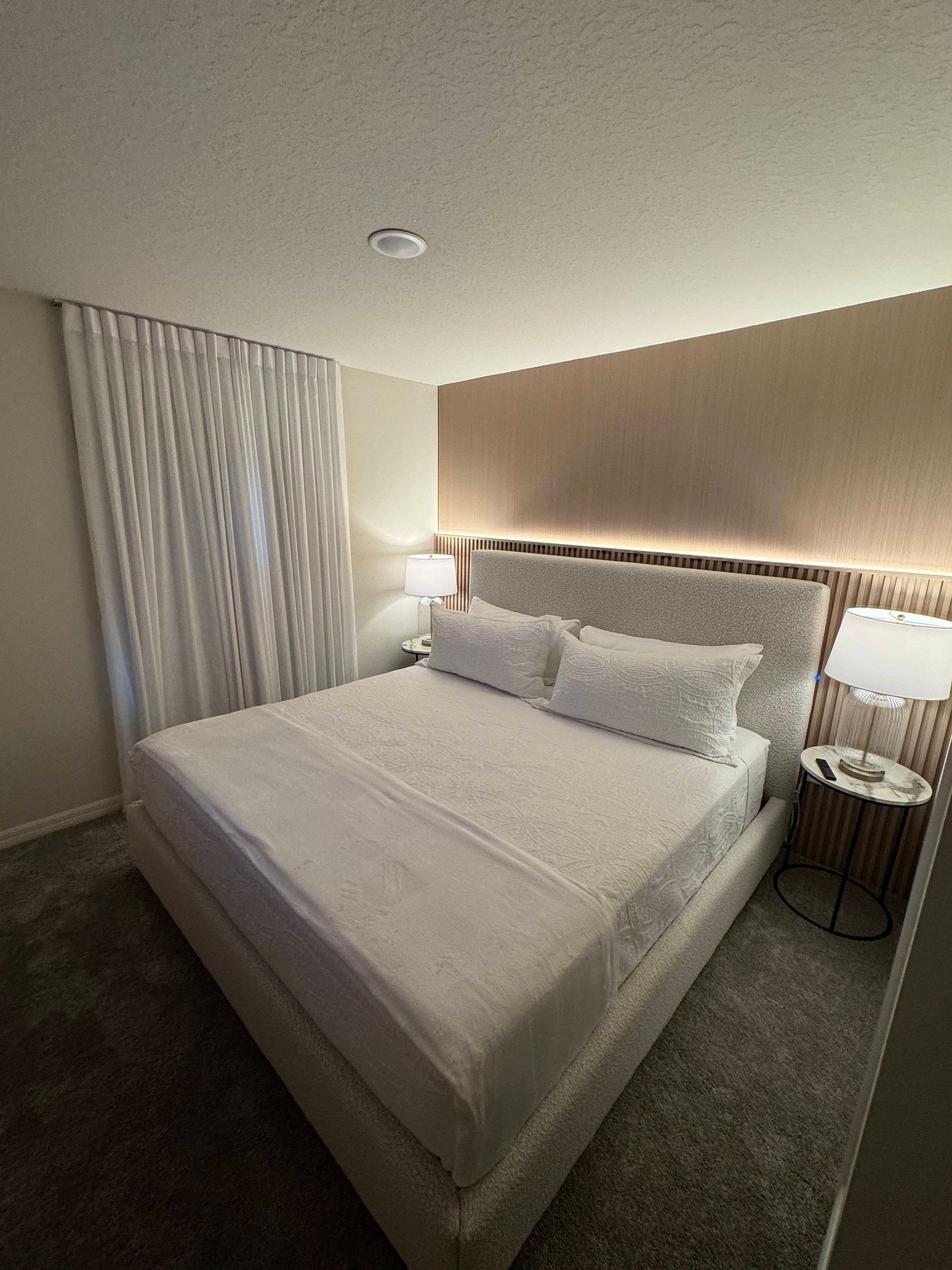 A hotel room with a large bed, two bedside tables, a headboard, a window with curtains, and a ceiling light.