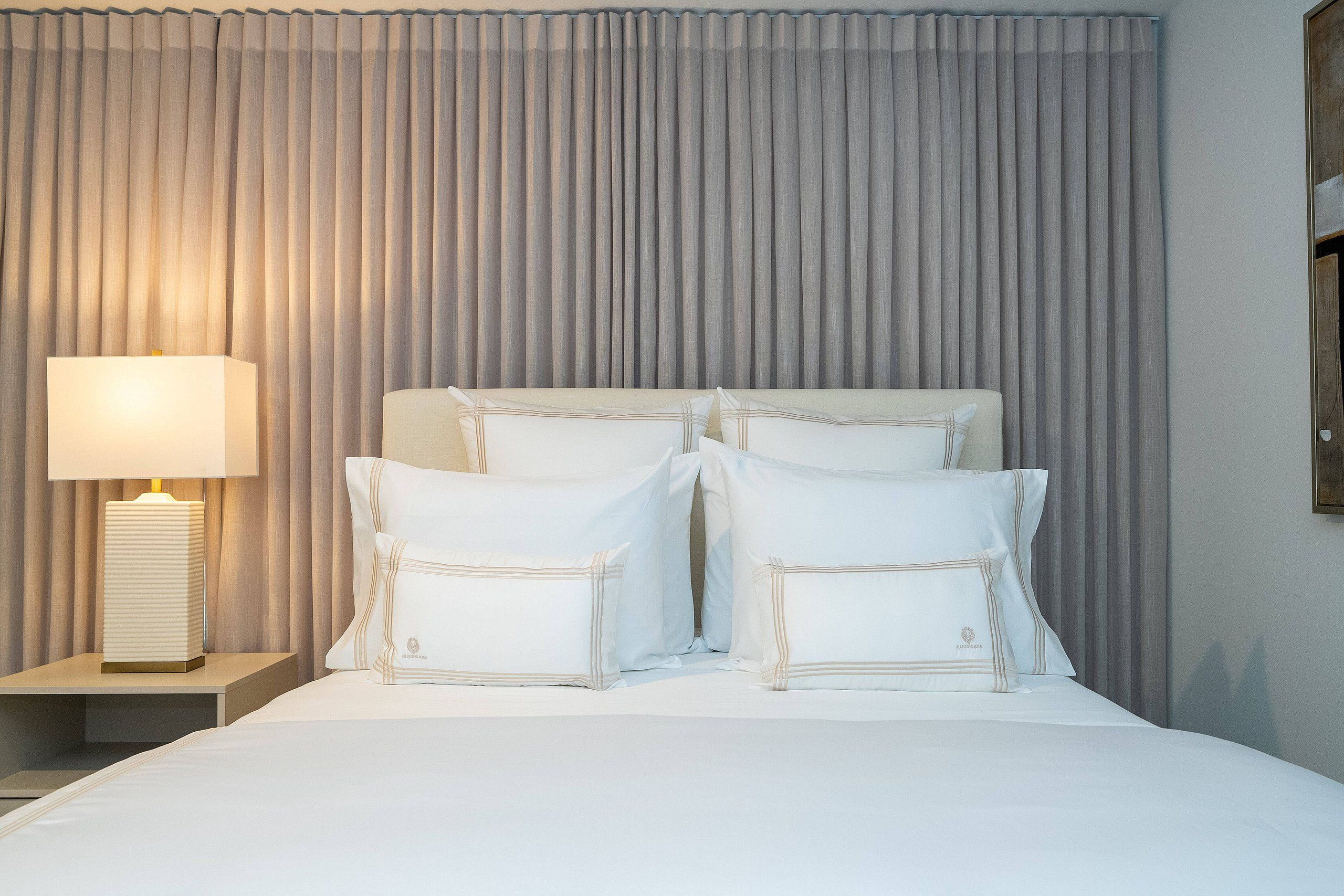 Una cama bien hecha con ropa blanca y almohadas, una lámpara de noche y un cortinaje de fondo.