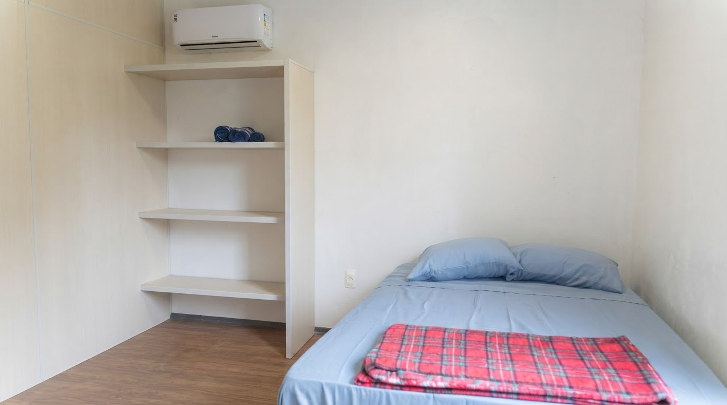 Una habitación pequeña y poco amoblada con una cama, una estantería y un aire acondicionado de pared.