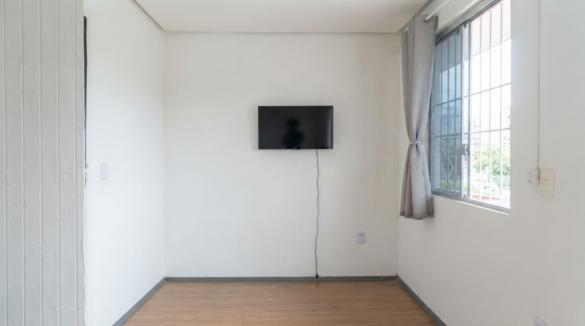 Una habitación con un televisor negro instalado en la pared, una ventana con cortinas y suelo de madera.