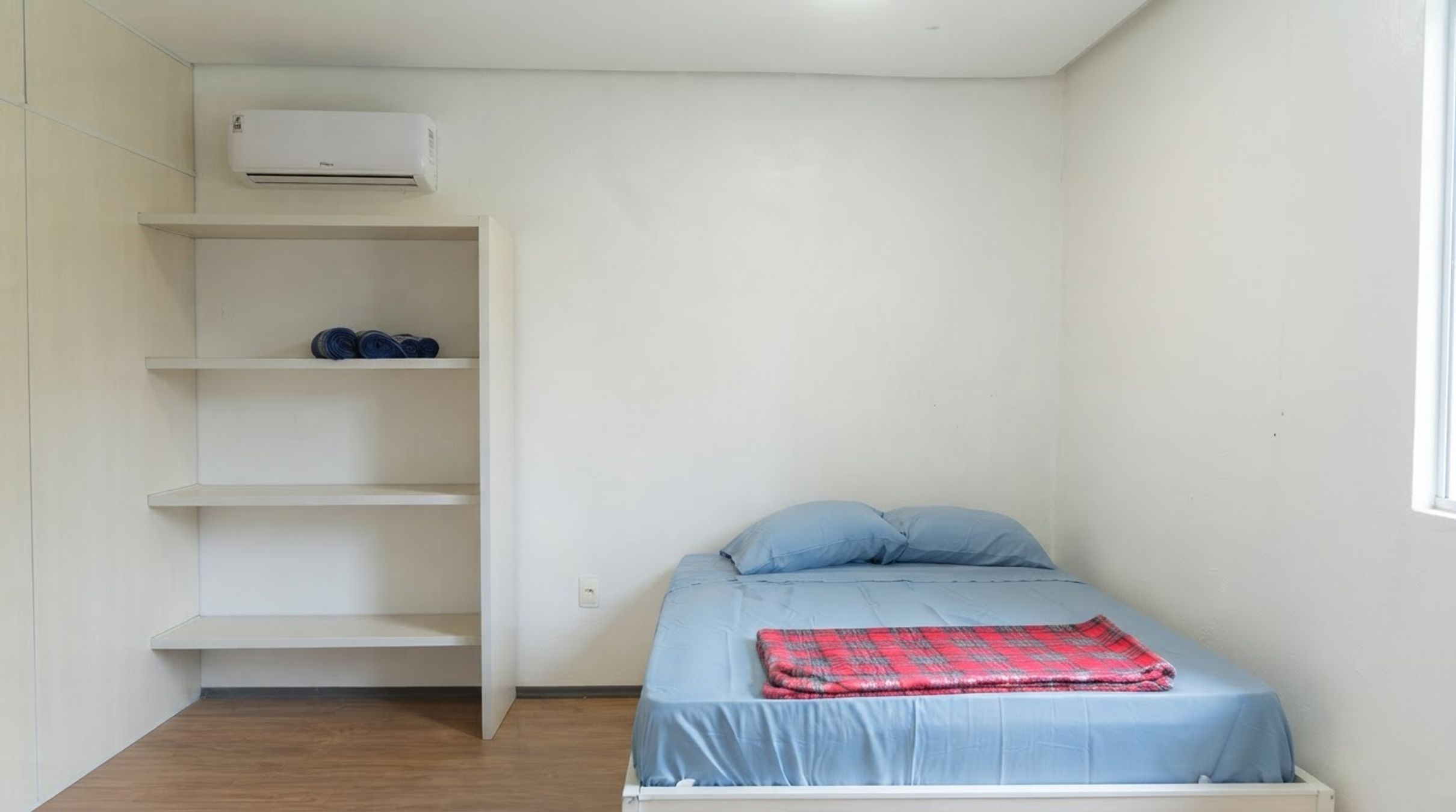 A small, single-bed room with a white air conditioning unit, a wooden floor, and a window on the right.