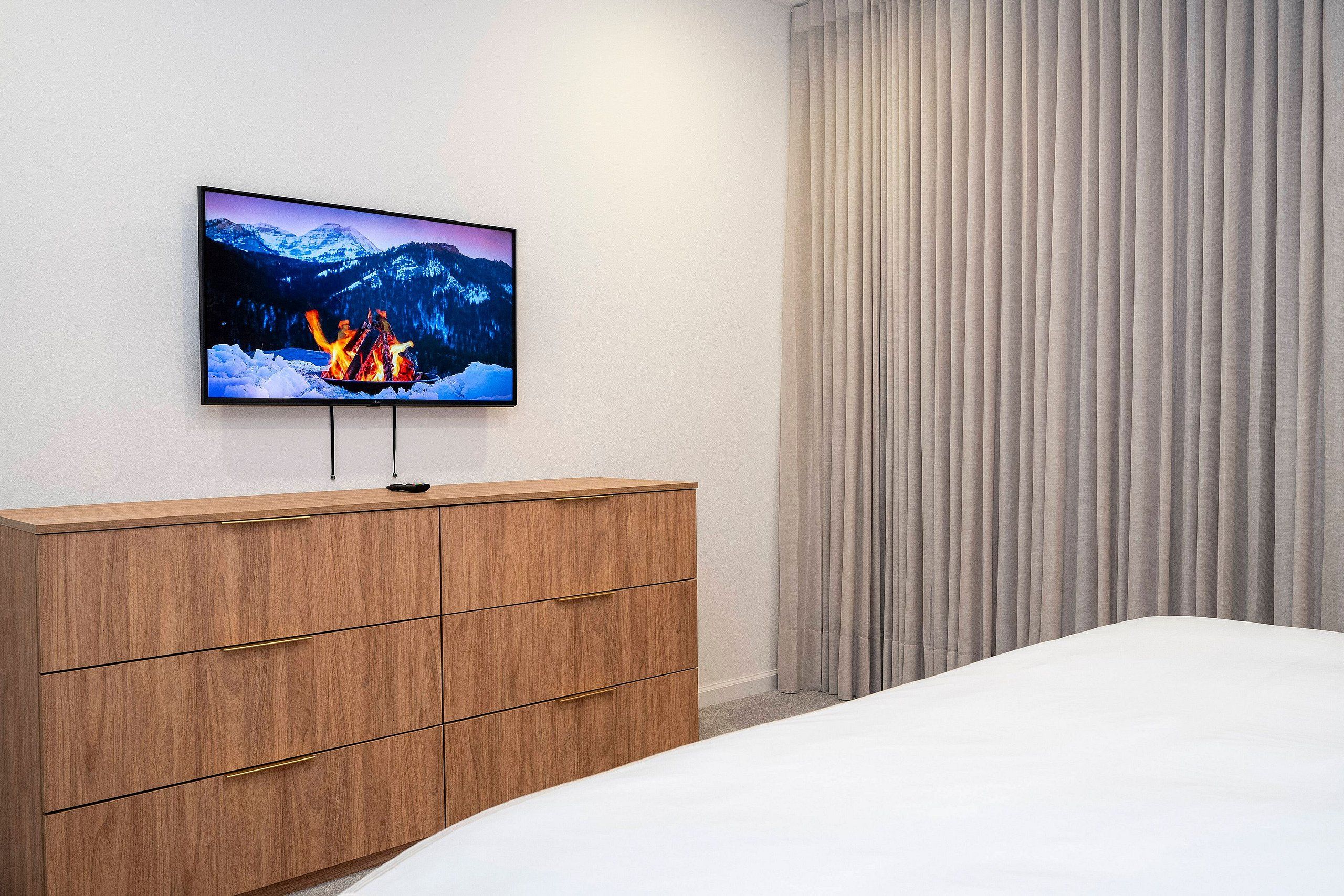 A bedroom with a bed, a wooden dresser, a mounted TV displaying a mountain scene, and a window with vertical blinds.