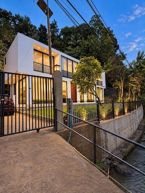 Une maison moderne à deux étages, dotée d’un balcon, d’une allée privée fermée par un portail et d’un petit ruisseau qui la borde.