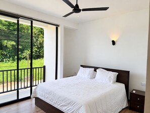 Ein Schlafzimmer mit einem großen Bett, einem Deckenventilator, einem Nachttisch und Blick auf Grün durch eine Schiebetür.