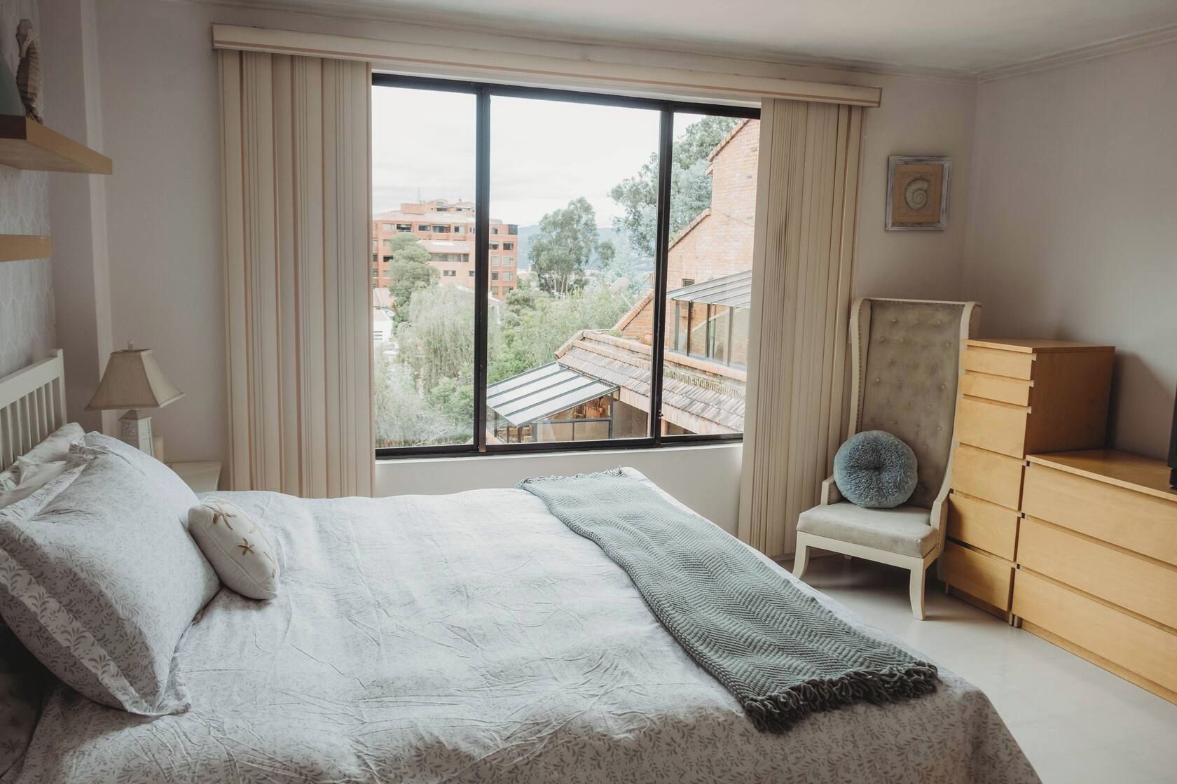 A bedroom with a large window, a bed with pillows, a chair, and a dresser.