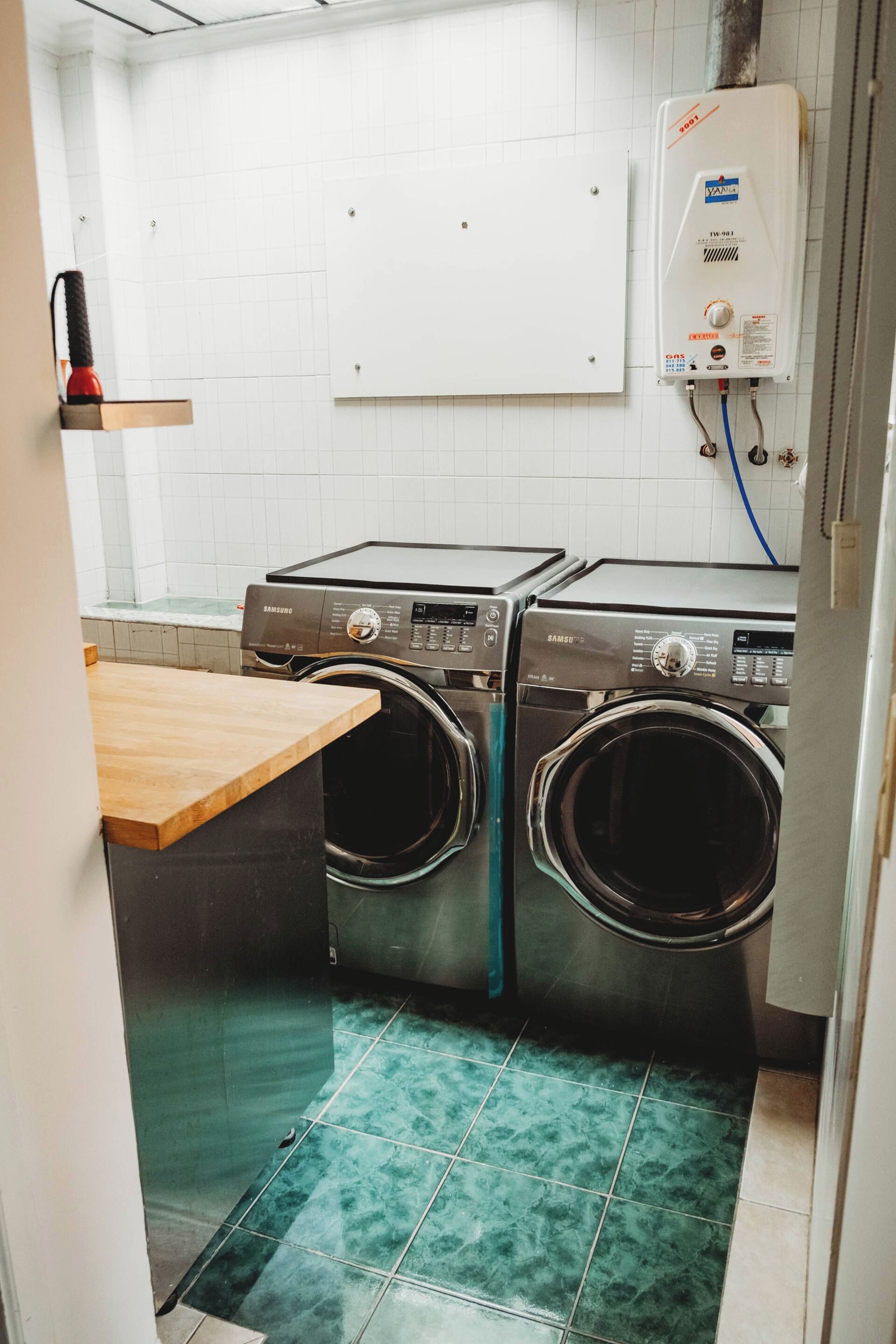 A compact laundry room with two front-loading washing machines, a wall-mounted heater, and a wooden counter.