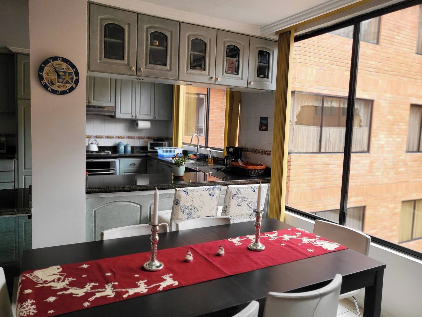 A dining area with a table set for two, a kitchen in the background, and a view of a brick building through a window.