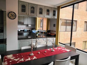 Un comedor con una mesa lista para dos, una cocina al fondo y vista a un edificio de ladrillo a través de una ventana.