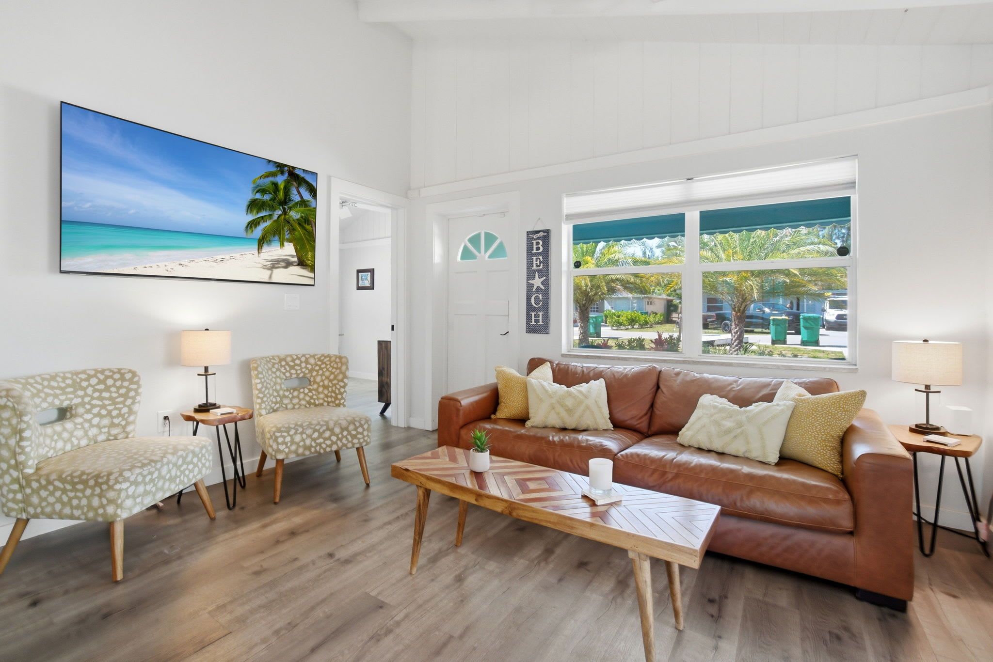 Un salon avec un canapé en cuir marron, des fauteuils à motifs, une table basse et un grand tableau sur le thème de la plage accroché au mur.