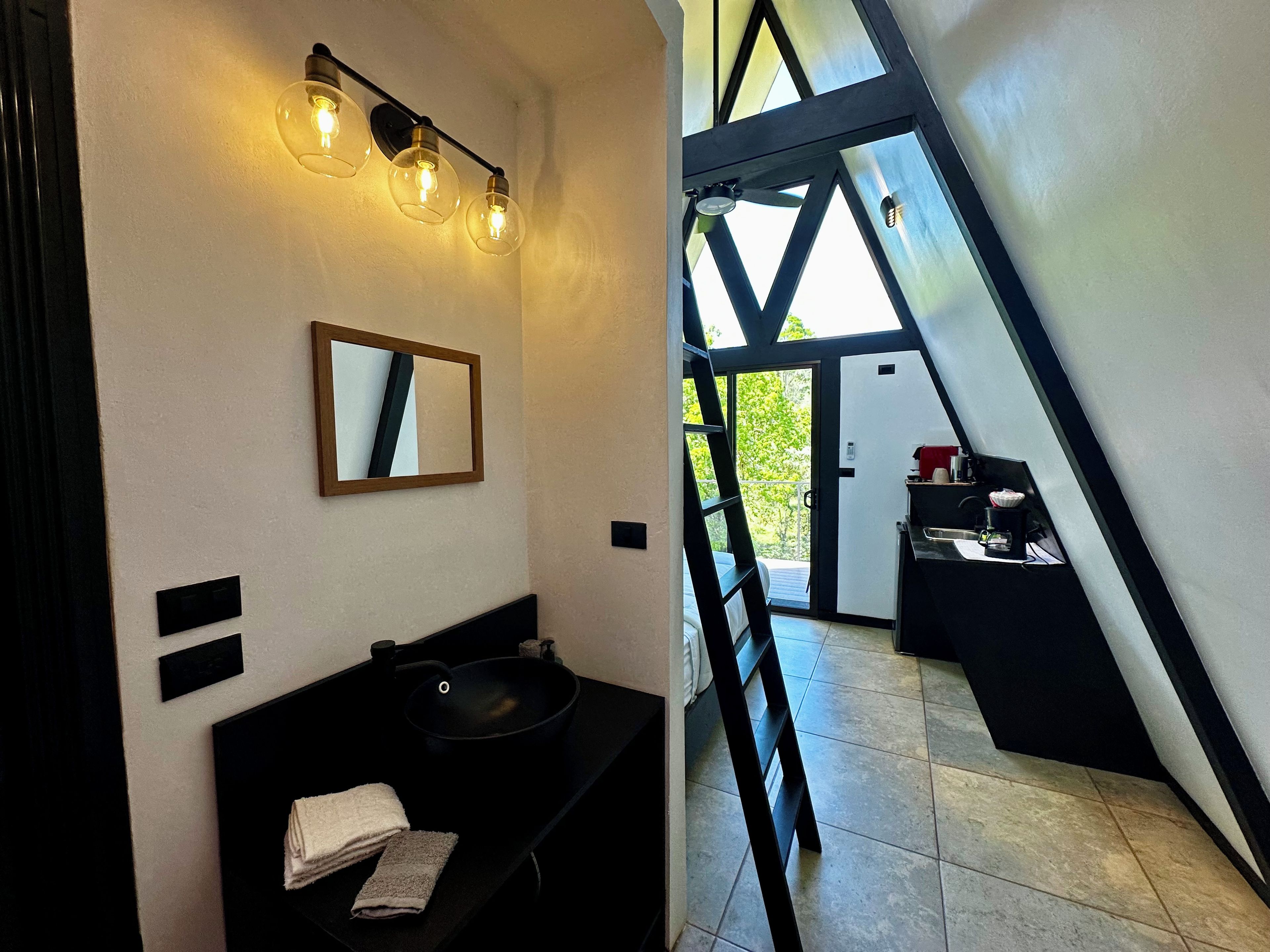 A modern bathroom with a black sink, a mirror, and a towel. There is a staircase leading to a balcony with a view of greenery.