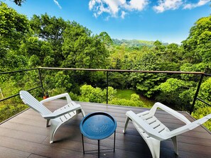 Eine Terrasse mit zwei weißen Liegestühlen und einem kleinen runden Tisch, mit Blick auf einen üppigen grünen Wald.