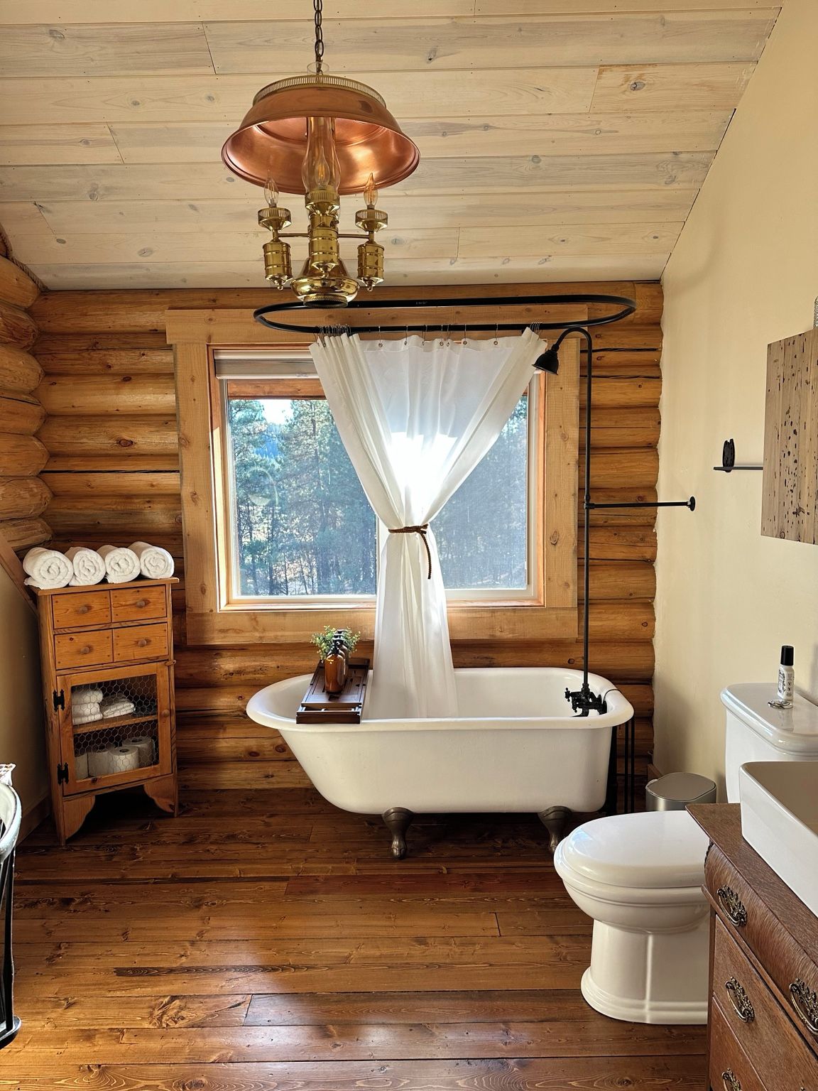 A log cabin bathroom with a freestanding bathtub, a white toilet, and a wooden cabinet with towels.
