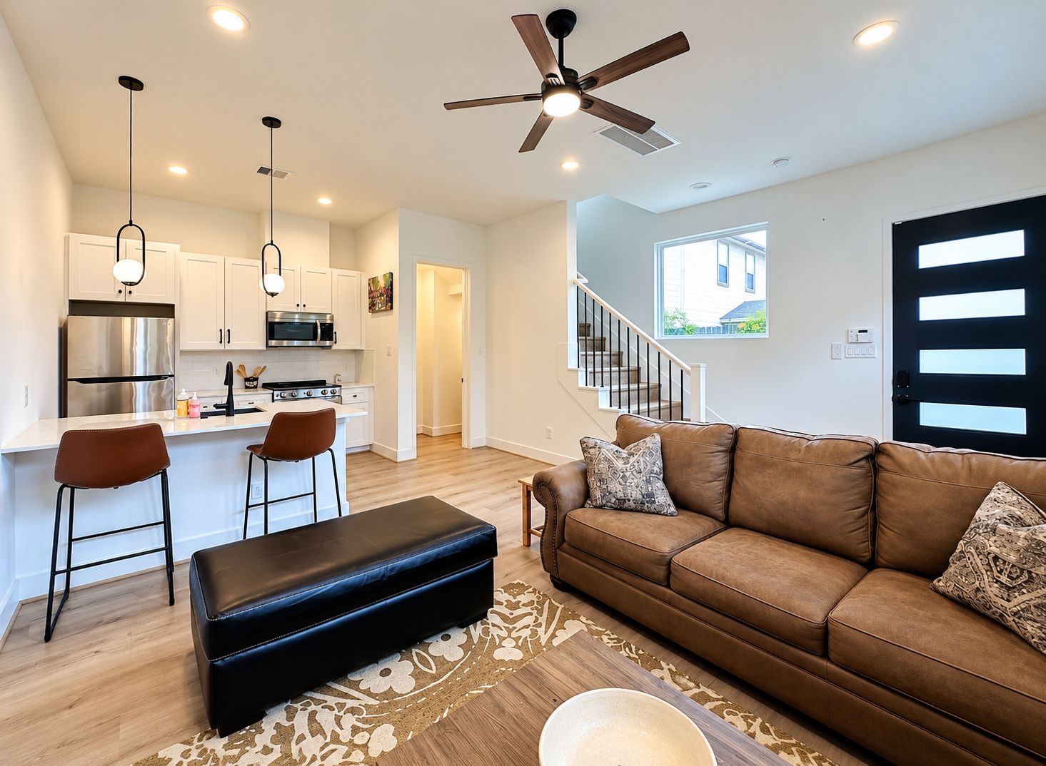 A modern kitchen with a central island, a dining area with bar stools, a brown leather sofa, and a ceiling fan.