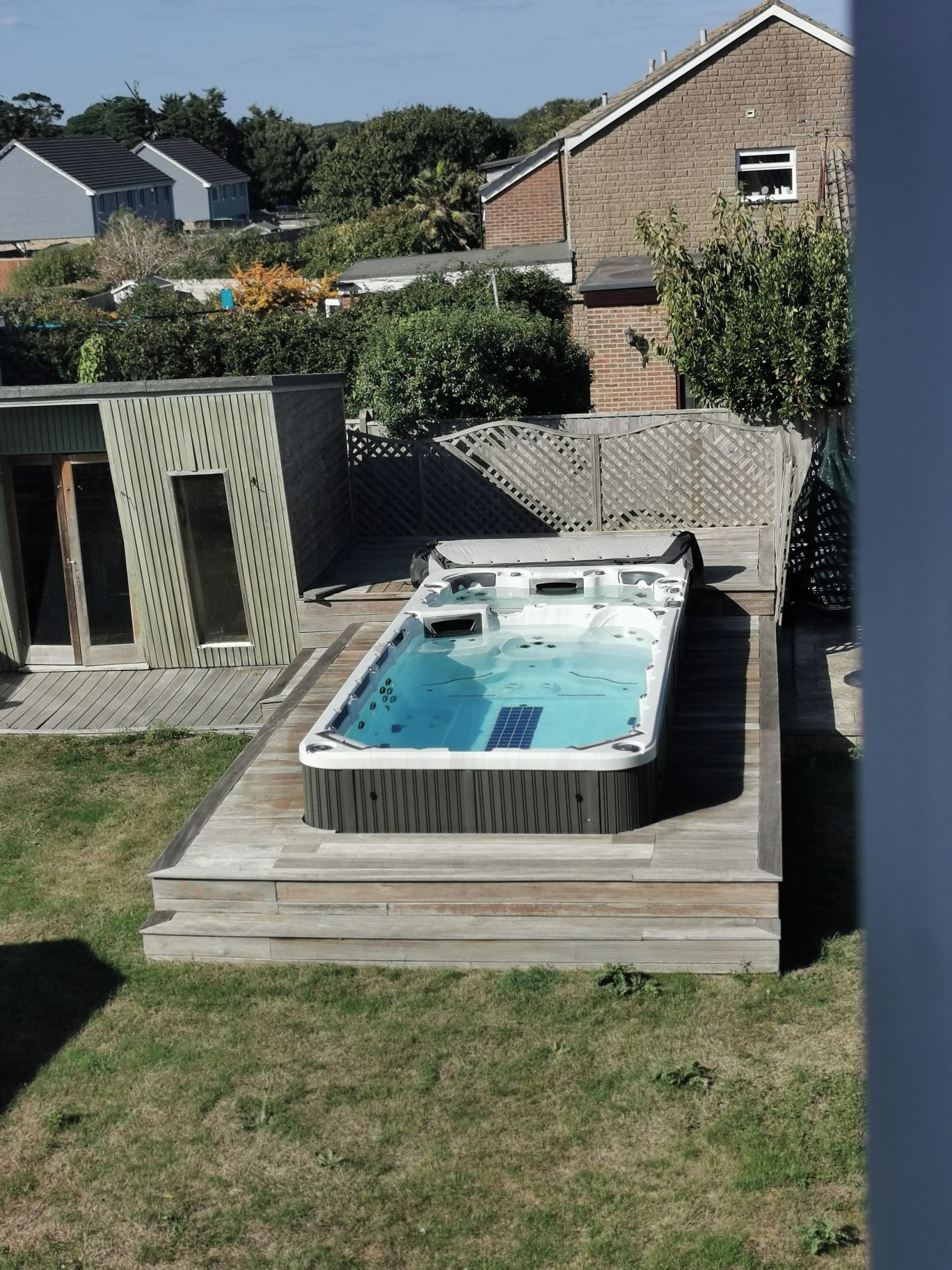 A rectangular hot tub with a built-in heater, surrounded by a wooden deck, set in a backyard with a brick house and greenery in the background.
