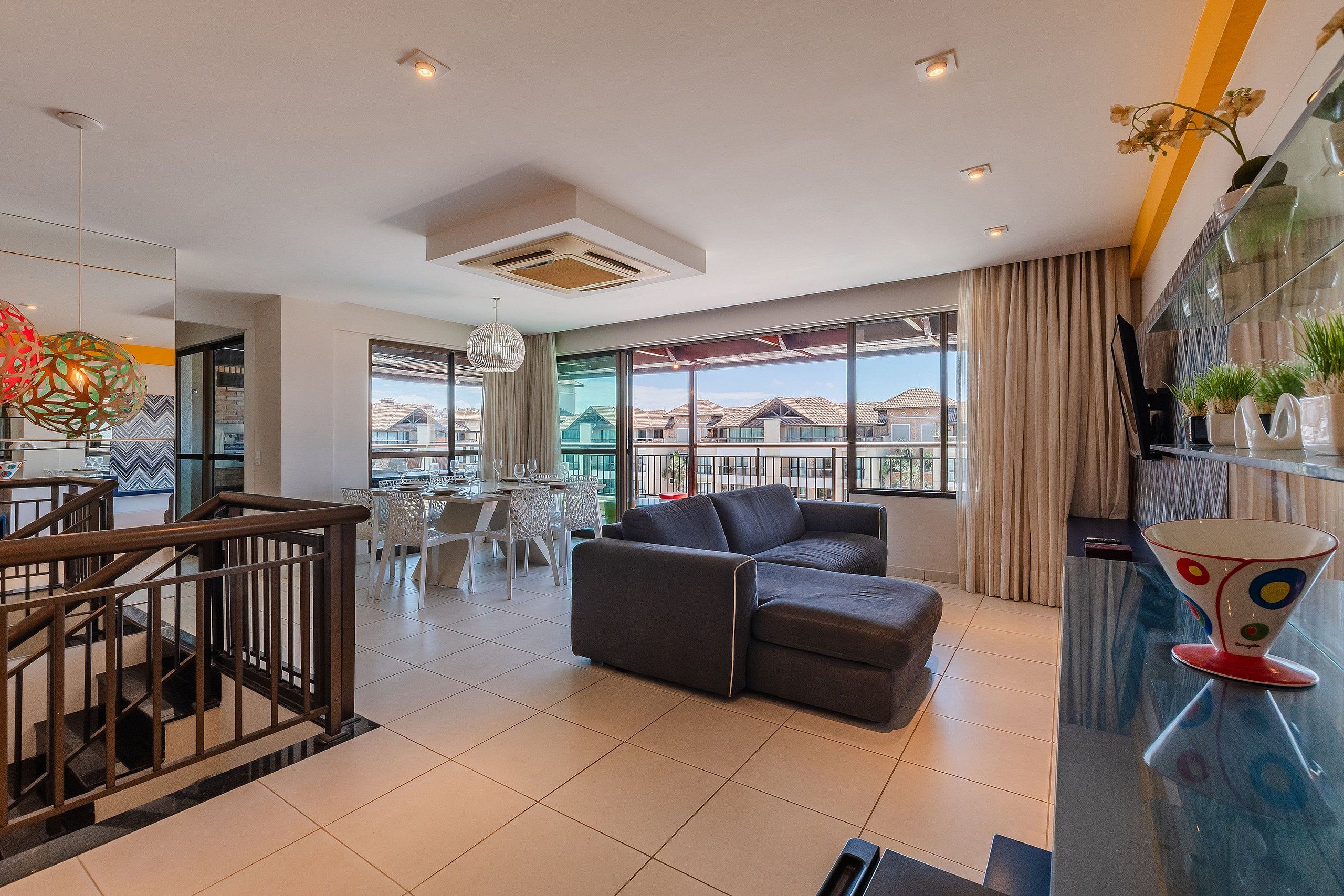 Uma sala de estar moderna com sofá, área de jantar e grandes janelas com vista para os edifícios ao redor.