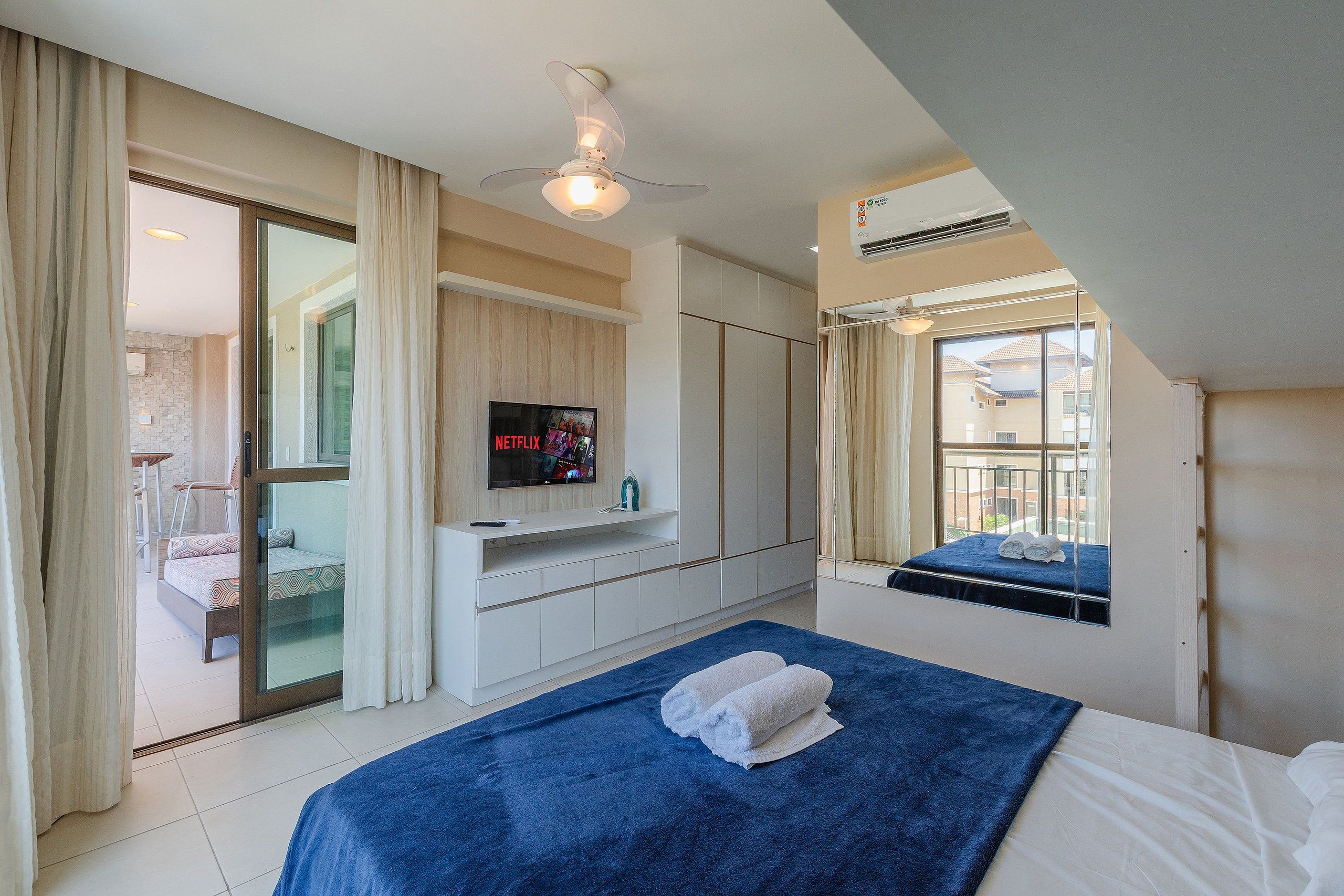 Quarto de hotel com cama, TV fixada na parede, ventoinha no teto e janela com vista para edifícios.