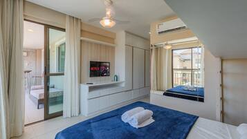 Quarto de hotel com cama, TV fixada na parede, ventoinha no teto e janela com vista para edifícios.
