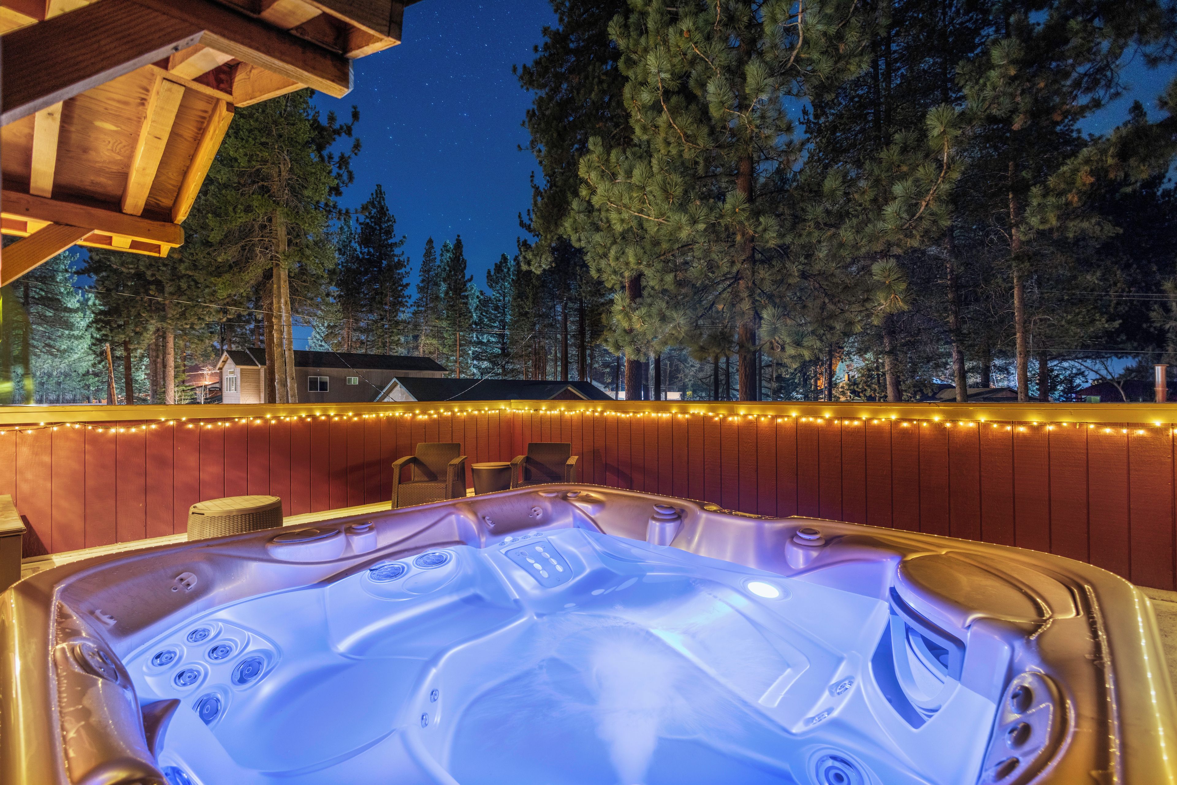 A hot tub with a wooden deck and a string of lights, surrounded by trees and a house in the background.