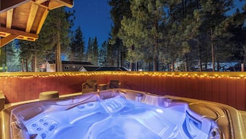 A hot tub with a wooden deck and a string of lights, surrounded by trees and a house in the background.