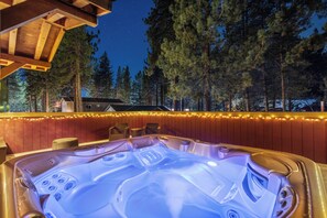 Un jacuzzi con terraza de madera y guirnalda de luces, rodeado de árboles y una casa al fondo.