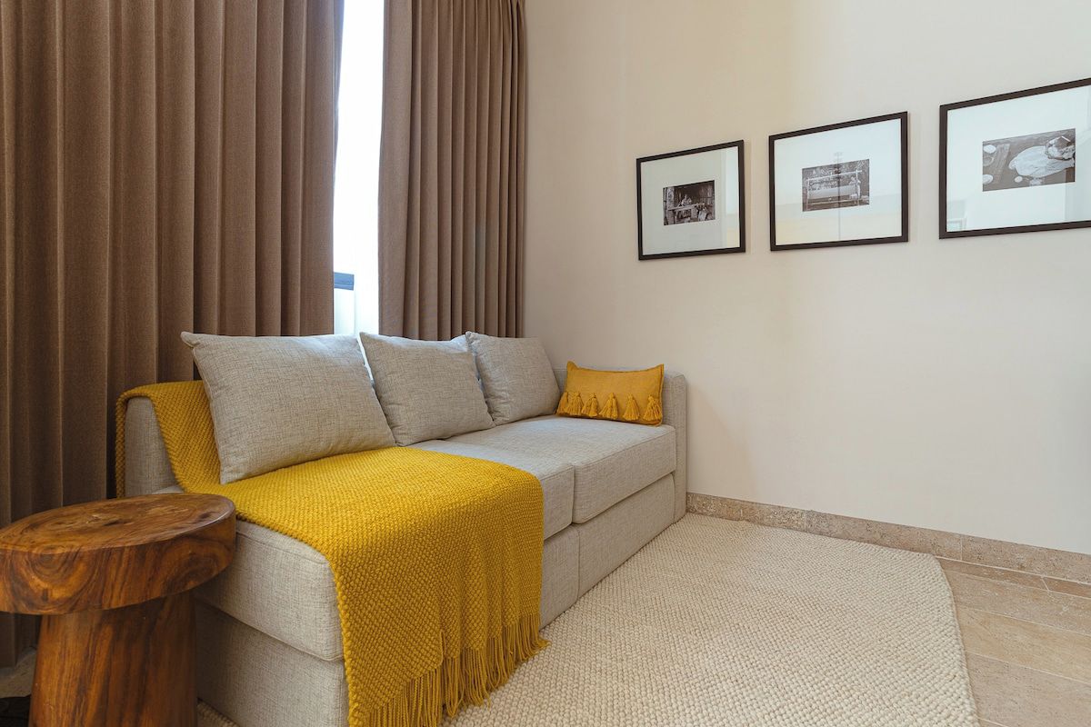 Un soggiorno con un divano, una coperta gialla, un piccolo tavolino in legno e un tappeto beige. Tre quadri sono appesi al muro.