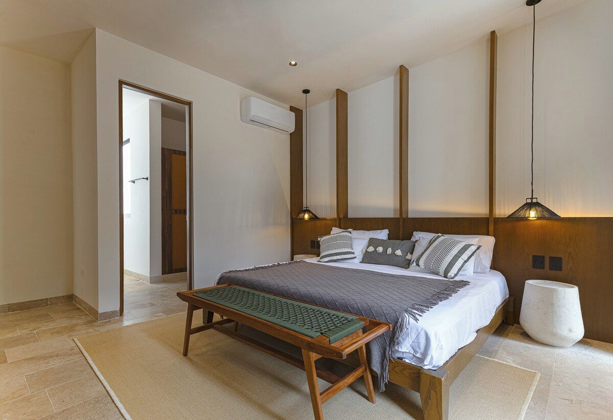 Une chambre à coucher comprenant un lit, des tables de chevet, un banc, un climatiseur fixé au mur et un luminaire suspendu.