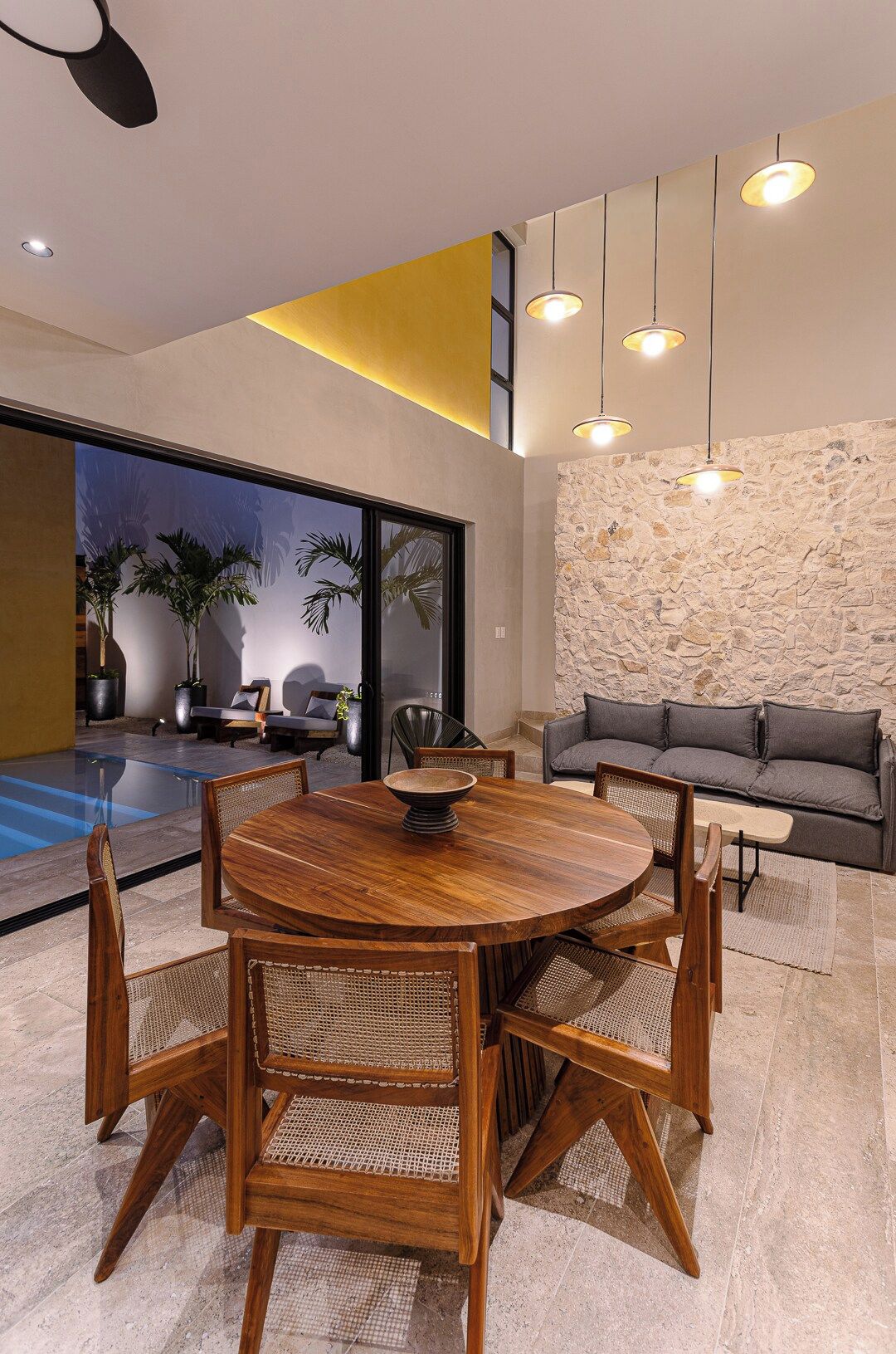 Un espace de repas moderne avec une table ronde en bois et des chaises en osier, un mur d’accent en pierre et un canapé gris.