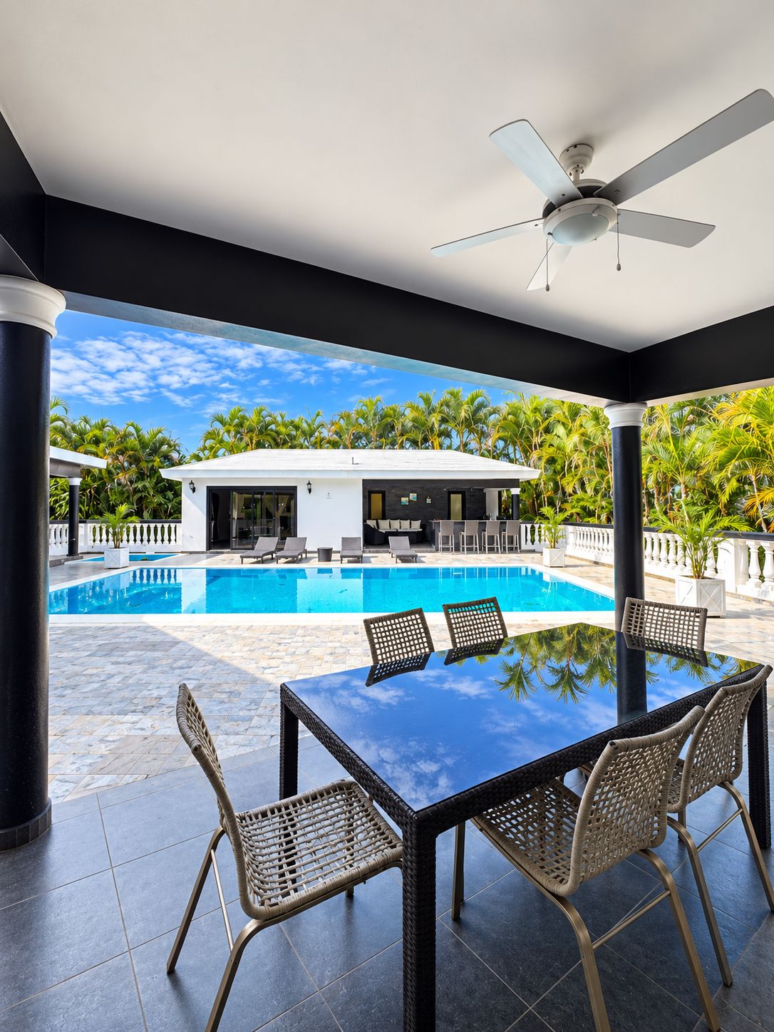 Un espace aménagé au bord de la piscine, avec une table et des chaises, un ventilateur de plafond et une vue sur un bâtiment doté d’une piscine.