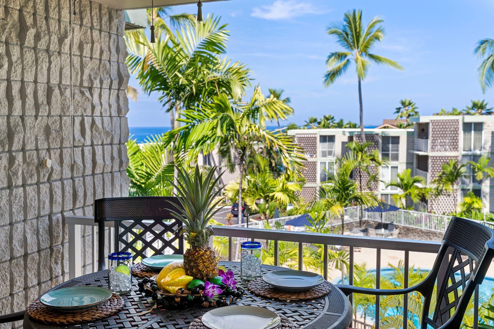 A balcony with a table set for dining, surrounded by tropical plants and overlooking a pool and hotel buildings.