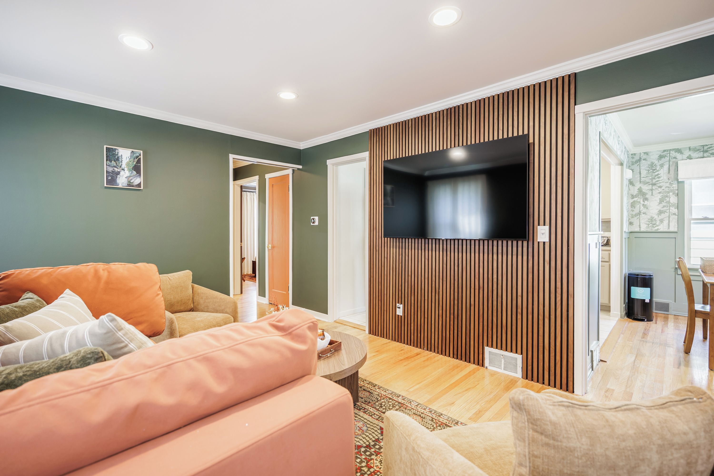 Una sala moderna con un televisor de pantalla plana montado en una pared de paneles de madera, un sofá con cojines, una mesa de centro y una zona de comedor al fondo.