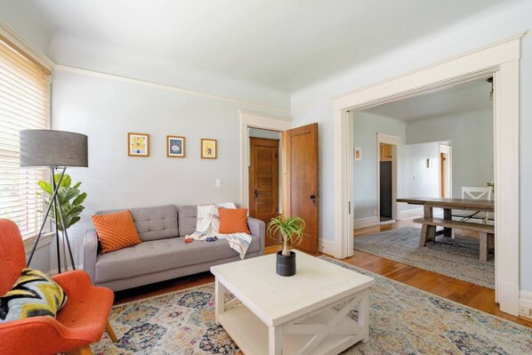 A living room with a sofa, a coffee table, a lamp, and a dining area with a table and chairs. - 1906 Craftsman • 5 Queen Beds • Sleeps 10 (Portland)
