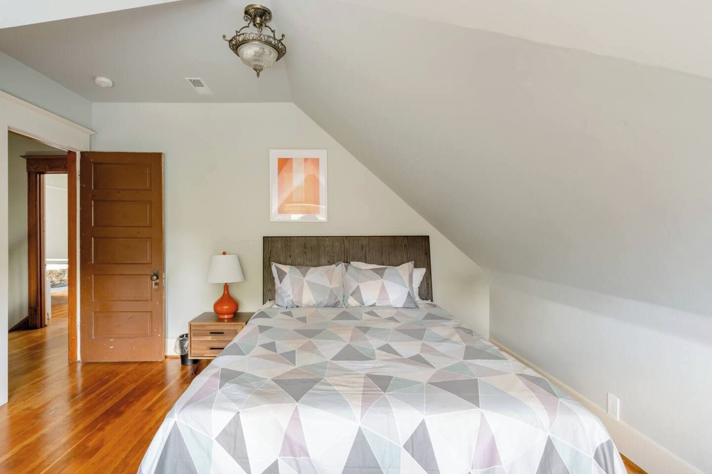 Une chambre avec un plafond en pente, un lit recouvert d’une couvre-lit à motifs géométriques, une table de chevet avec une lampe, et une porte donnant sur une autre pièce.