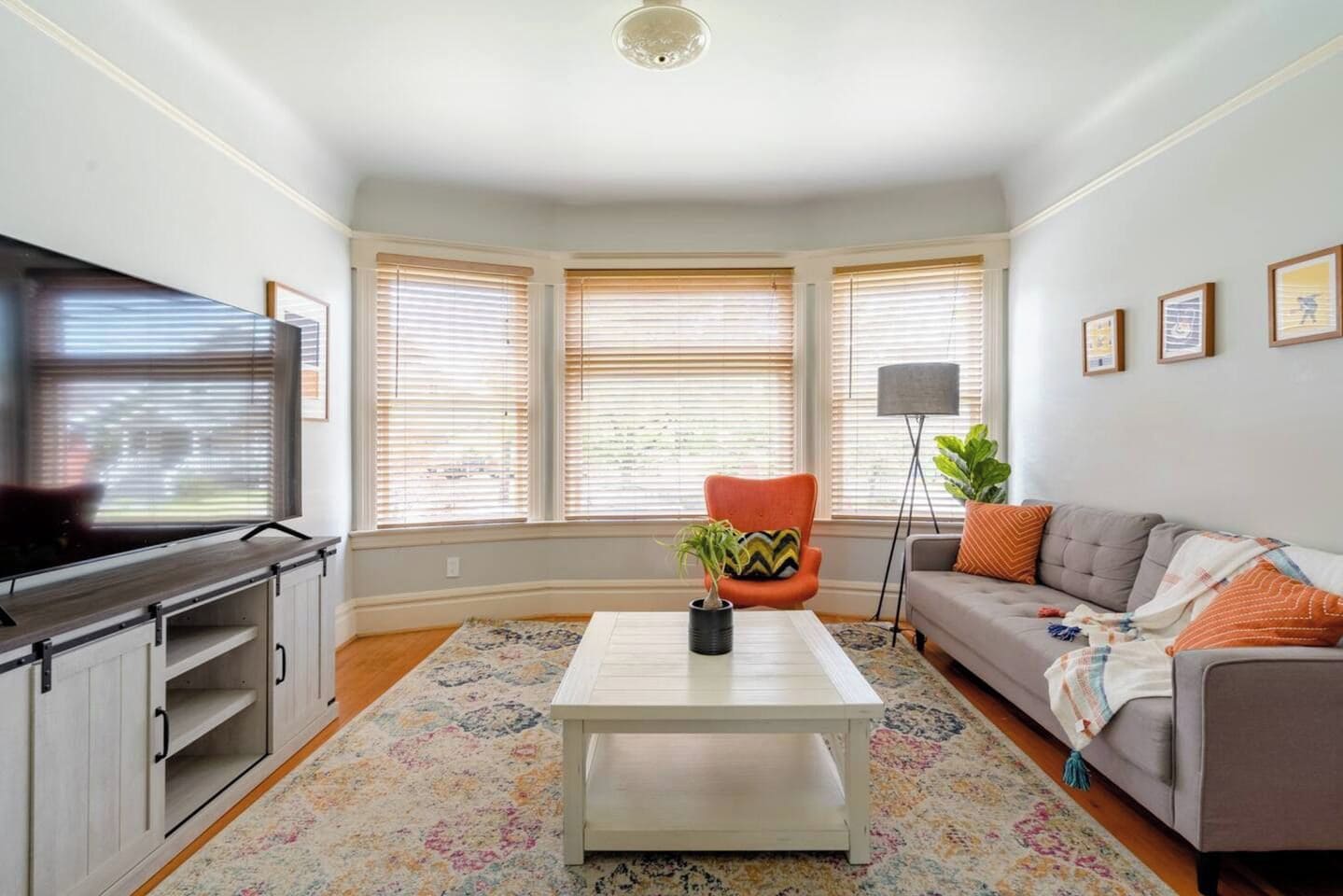 A living room with a television, a sofa, a coffee table, and a chair. There are three windows with blinds, a floor lamp, and a plant.