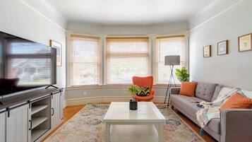 A living room with a television, a sofa, a coffee table, and a chair. There are three windows with blinds, a floor lamp, and a plant.