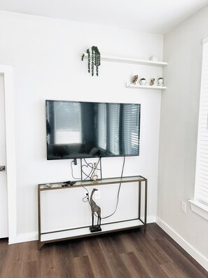 Ein modernes Wohnzimmer mit einem an der Wand befestigten Flachbildfernseher, einem Konsolentisch mit Dekoration und einem Topf mit Pflanze auf einem weißen Regal.