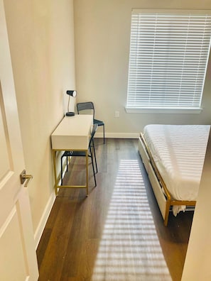Ein kleines Zimmer mit einem Bett, einem Schreibtisch und einem Stuhl. Es gibt ein Fenster mit Jalousien, und der Boden ist aus Holz.
