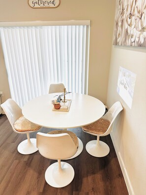 Una mesa redonda para comer con cuatro sillas, una pared blanca con un cuadro enmarcado y una ventana con persianas verticales.
