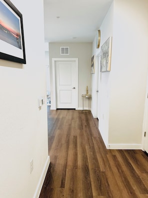 Un pasillo con suelo de madera, paredes blancas, una puerta al fondo y una pequeña mesa con un objeto decorativo.