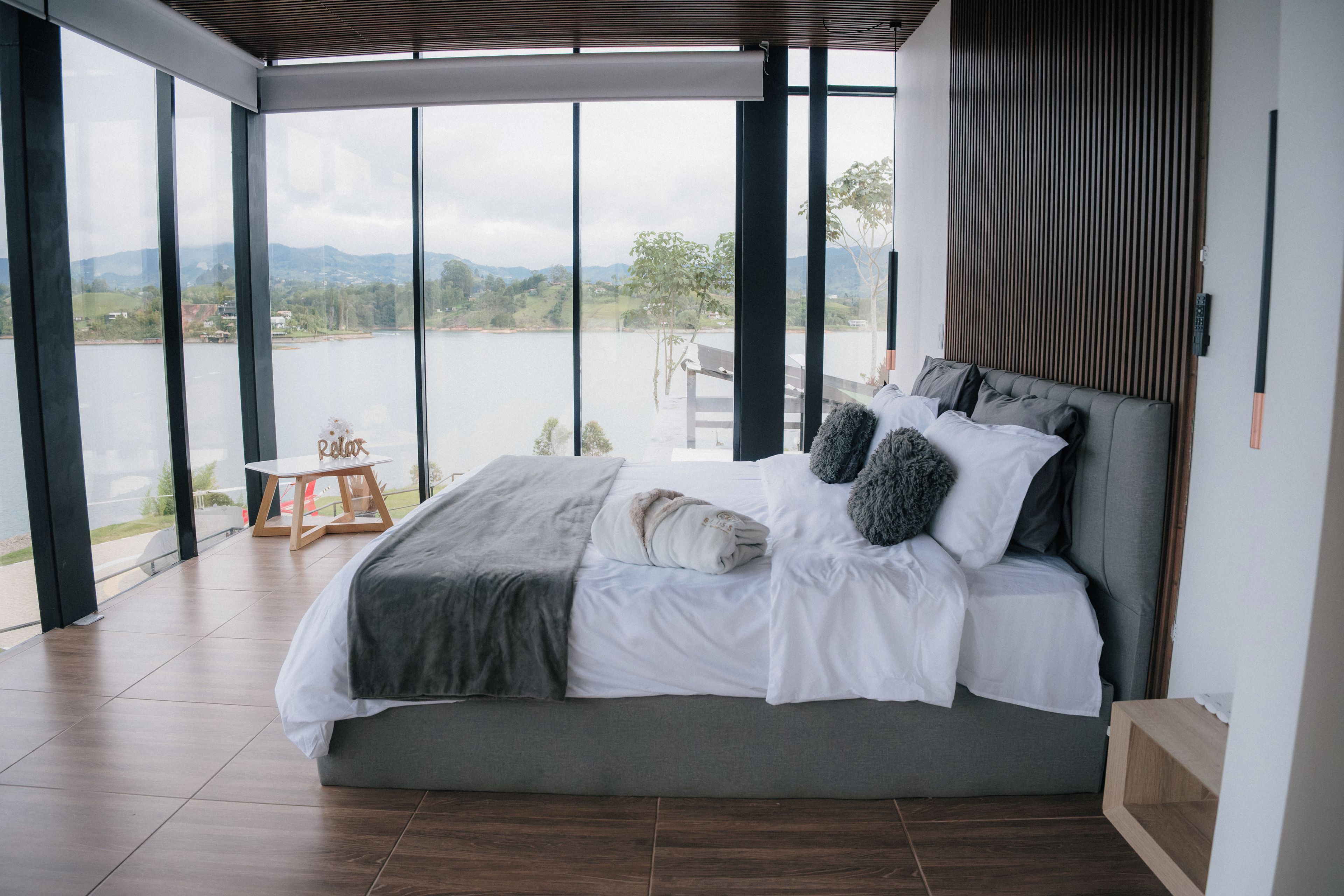 Un dormitorio moderno con una cama grande, vista a un lago y una pequeña mesa con una silla.