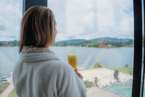 A person holding a glass of orange juice, looking out at a lake with mountains in the distance. - Bliss Club (El Peñol)