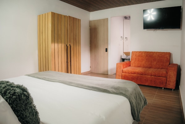A bedroom with a bed, an orange sofa, a wooden wardrobe, and a flat-screen TV on the wall. - Bliss Club (El Peñol)