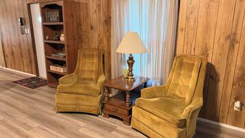 Une pièce aux murs lambrés, avec deux fauteuils, une petite table sur laquelle se trouve une lampe, une bibliothèque et un téléphone.