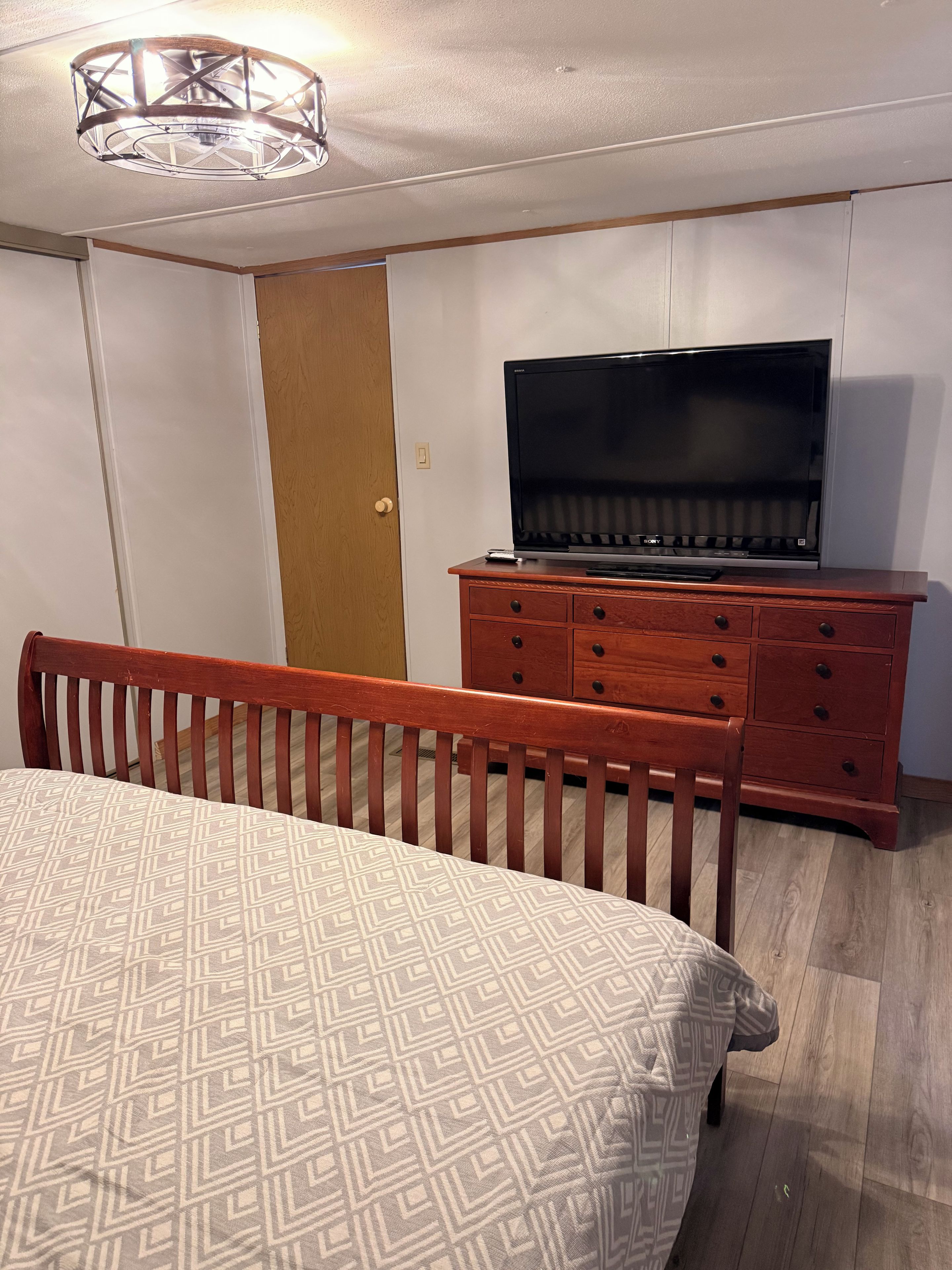 Un dormitorio con una cama, un aparador de madera con un televisor, una puerta y una lámpara en el techo.