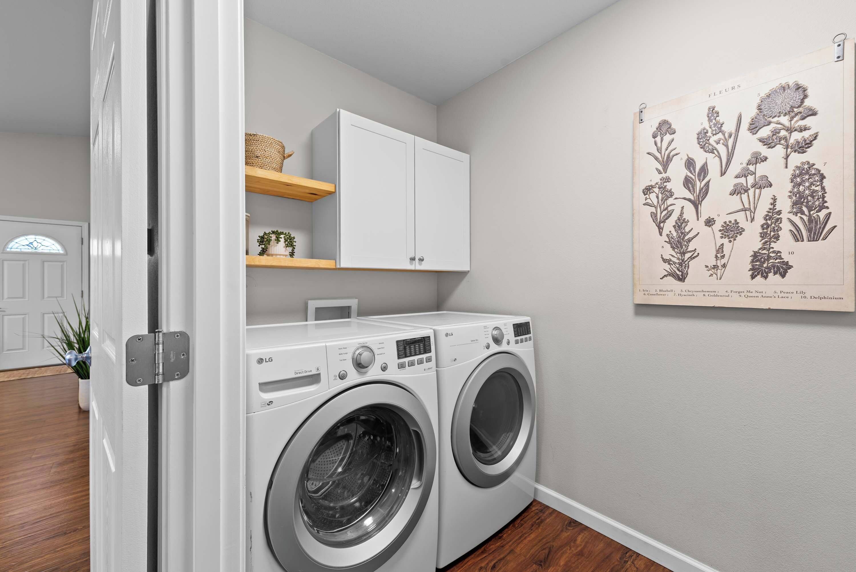 A laundry room with two front-loading LG washers, a white cabinet above, and a botanical illustration on the wall.