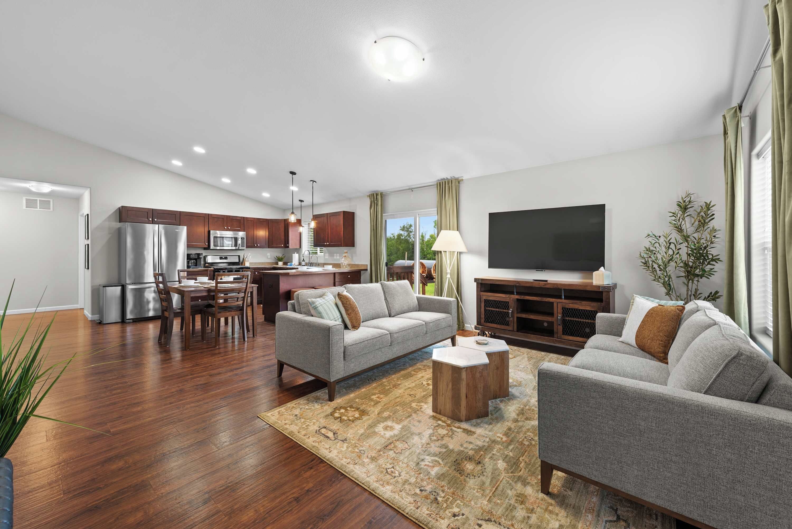 A modern living room with a flat-screen TV, grey sofas, a coffee table, and a kitchen area with a refrigerator, microwave, and dining set.