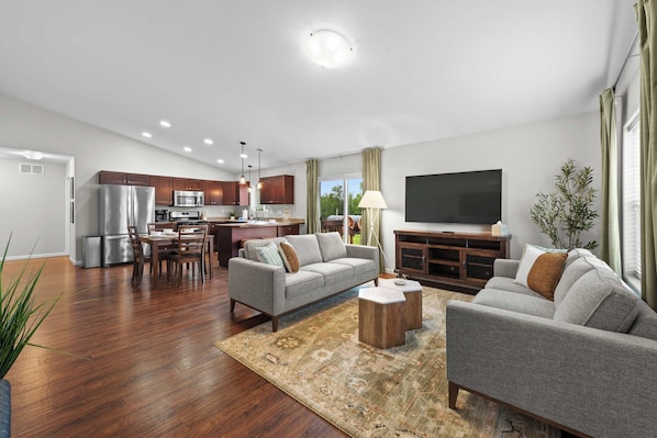 A modern living room with a flat-screen TV, grey sofas, a coffee table, and a kitchen area with a refrigerator, microwave, and dining set. - Quiet 3BR Getaway w/ Full Kitchen & Laundry! (Wasilla)