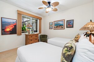 Habitación de hotel con dos camas, un ventilador de techo, una ventana con cortinas, un aparador de madera, una silla y cuadros en la pared.