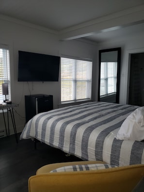 A bedroom with a bed, a television, a desk, and a window with blinds. - Rose Castle Inn & Café. Beautiful, breathtaking views of the ocean. (Bell Island)
